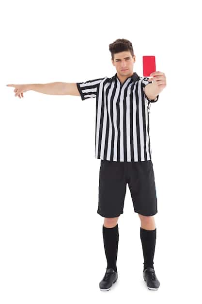 A referee gesturing a VAR check, drawing a rectangle in the air, with a soccer pitch and players in the background.