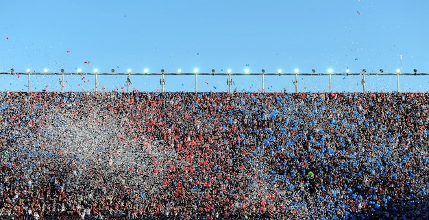 A vibrant crowd of US soccer fans, predominantly in red, white, and blue, cheering passionately at a large outdoor public viewing party for a World Cup match, with a giant screen showing the game.