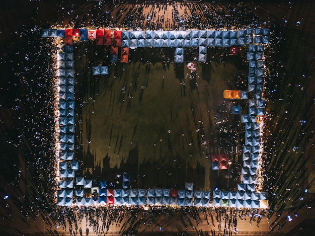 An aerial view of a World Cup fan zone at night, showing a densely packed crowd of people in front of a giant screen. The scene is illuminated by bright lights and the glow of the screen, with food stalls and sponsor tents lining the perimeter. The atmosphere is festive and energetic, with people cheering, dancing, and celebrating together at this World Cup Fan Zones: Where Will the Biggest Public Viewing Parties Be Held Across the US in 2026?.